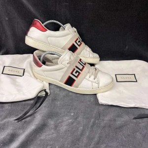 Vita sneakers från Gucci med röd detalj - Säljer ett par vita sneakers från Gucci med breda elastiska band över foten där det står 'GUCCI' i svart och rött. Skorna har röda detaljer på hälen och klassisk rund tå. Snygg och sportig design med tydlig logga.