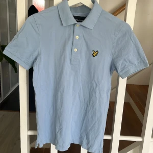 Ljusblå pikétröja från Lyle & Scott - Säljer en ljusblå pikétröja från Lyle & Scott med klassisk krage och två knappar framtill. Tröjan har korta ärmar och den ikoniska gula loggan broderad på bröstet. Perfekt för en stilren och avslappnad look.