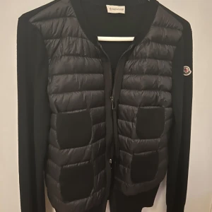 Moncler Cardigan  - Säljer min svarta cardiganjacka från Moncler med quiltade partier framtill och fyra fickor. Jackan har långärmade stickade ärmar och dragkedja framtill. Klassisk Moncler-logga på ärmen och rund halsringning. Sparsamt använd och väldigt bra skick, köpt på Luisaviaroma. 