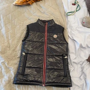 Svart dunväst från Moncler - Snygg svart dunväst från Moncler med glansig finish och klassisk Moncler-logga på bröstet. Västen har två fickor med spännen och en röd, vit och blå randig dragkedja framtill. Perfekt för lager-på-lager under kyliga dagar.