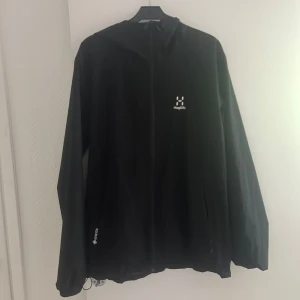 Svart vindjacka från Haglöfs - Säljer en svart vindjacka från Haglöfs med huva och dragkedja framtill. Jackan är tillverkad i syntetmaterial och har diskret logga på bröstet. Perfekt för utomhusaktiviteter och träning.