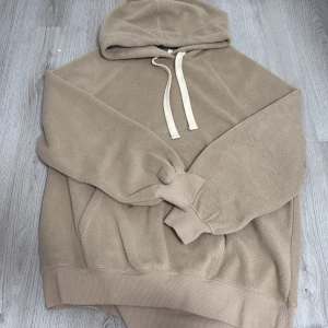 Beige fleece hoodie från H&M - Mysig beige hoodie från H&M i fleece-material. Tröjan har huva med dragsko och breda muddar vid ärmslut och nederkant. Perfekt för en avslappnad stil och extra värme under kyliga dagar.
