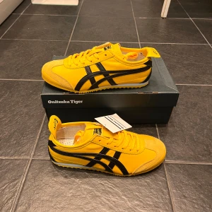 Gula Onitsuka Tiger Mexico 66 sneakers - Säljer ett par ikoniska Onitsuka Tiger Mexico 66 sneakers i gul färg med svarta detaljer. Skorna har klassisk låg profil, snörning och är tillverkade i syntetmaterial och skinn. Perfekta för dig som vill sticka ut med färg på fötterna!