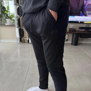Svarta mjukisbyxor från BOSS - Svarta mjukisbyxor från BOSS med logga på vänster lår. Byxorna har en slim passform och är tillverkade i mjukt material, perfekt för en avslappnad stil. Riktigt sköna mjukisbrallor men komme ej till användning 