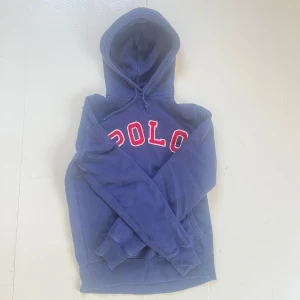 Blå hoodie från Polo Ralph Lauren - Mörkblå hoodie från Polo Ralph Lauren med stor röd och vit POLO-text på bröstet. Klassisk känguruficka framtill och justerbar huva. Perfekt för en avslappnad stil. Skicket är mycket bra, inga slitningar eller misfärggningar. Säljer pga för liten. Det är bara att höra av sig vid funderingar.