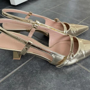 Guldiga pumps med remmar och klack - Säljer ett par eleganta pumps i guld med spetsig tå och två smala remmar över foten. Skorna har en låg klack och öppen häl, vilket ger en stilren och trendig look. Perfekta för att lyfta vilken outfit som helst. Endast använda 1 gång, strl 39 men passar mig som pendlar mellan 38/39. 