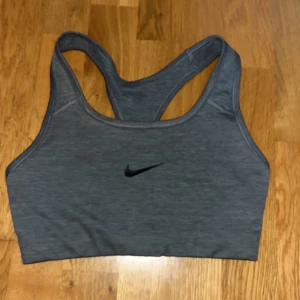 Grå sport-bh från Nike - Säljer en grå sport-bh från Nike med klassisk svart swoosh-logga framtill. Modellen har brottarrygg och är tillverkad i ett stretchigt material som passar perfekt för träning.