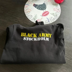 AIK hoodie - Svart hoodie med tryck 'BLACK ARMY STOCKHOLM' i gult och vitt på bröstet. Klassisk känguruficka framtill, huva med snörning och långa ärmar. Perfekt för dig som vill visa support för Black Army och AIK. Mjuk och skön, passar till chill eller matchdag.