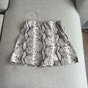 Ormmönstrad kortkjol från H&M XS - Snygg kortkjol från H&M i ormmönstrat tyg med beige, svart och grå toner. Kjolen har en liten slits framtill och bred resår i midjan för extra komfort. Materialet är mjukt och stretchigt med viskos och polyamid.