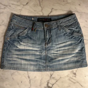 Blå lågmidjad jeanskjol från Musketeer Denim - Snygg blå lågmidjad jeanskjol från Musketeer Denim med slitna detaljer och klassiska fickor både fram och bak. Kjolen har bälteshällor, knapp och dragkedja framtill samt coola kontrastsömmar. Perfekt för dig som gillar en avslappnad och trendig stil.