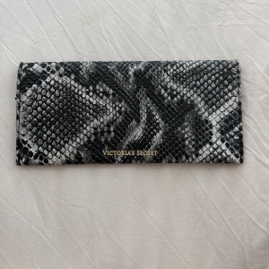 Plånbok i ormskinnsmönster Victoria's Secret - Snygg plånbok från Victoria's Secret i svartvitt ormskinnsmönster med guldfärgad logga och knapp. Plånboken har flera fack för kort och sedlar, och stängs med tryckknapp. Perfekt accessoar för dig som gillar djurmönster och stilren design.