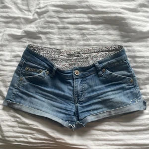 Lågmidjade jeansshorts - Snygga blå jeansshorts från Lonky Jeans med coola paljettdetaljer på bakfickorna och blommiga knappar. Shortsen har låg midja, uppvikta benslut och klassisk femficksdesign. Perfekta för sommaren och ger en avslappnad vibe. Man kan matcha shortsen med allt, man kan även välja om man vill vika upp shortsen så fickorna syns mera, eller vika ner så fickorna inte syns. Skriv om mer bilder eller funderinga💕