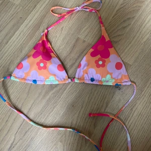 Blommig trekantsbikini i orange och rosa - Superfin trekantsbikini med stora blommor i rosa, lila, blått och grönt på en orange botten. Knytband i nacken och ryggen för perfekt passform. Materialet är mjukt och stretchigt i polyester och elastan. Perfekt för stranden eller poolen!