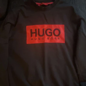 Svart tröja med röd HUGO BOSS-logga - Svart långärmad tröja från HUGO BOSS med stor röd logga framtill. Klassisk rund halsringning och relaxed fit. Perfekt för dig som gillar stilrena plagg med tydlig branding. Materialet känns mjukt och skönt mot huden.