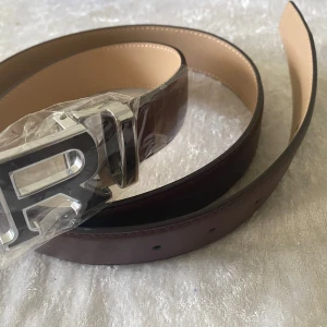 Brunt skärp med silverfärgat R-spänne - Snyggt brunt bälte i skinn med ett stort, silverfärgat R-spänne. Bältet har en stilren design och är perfekt för att lyfta vilken outfit som helst. Insidan är ljusbeige och sömmarna är diskreta för en clean look.