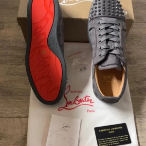 Grå mocka sneakers från Louboutin - Snygga grå sneakers från Christian Louboutin i mocka med ikoniska röda sulor och coola nitar på tån. Skorna har snörning och en rund tå, perfekt för dig som vill sticka ut med exklusiv streetstyle.