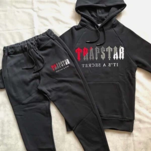 Trapstar tracksuit med tryck - Svart tracksuit från Trapstar med broderad logga i rött, grått och vitt på. Byxorna har dragsko i midjan och är tillverkade i mjukt bomullsmaterial. Perfekta för en chill och street-inspirerad look.