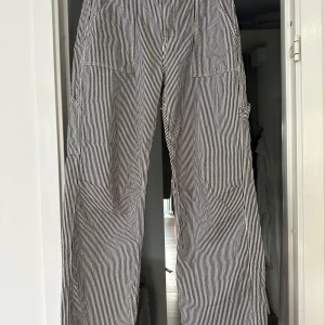 Randiga byxor i bomull - Säljer ett par svartvita cargopants med smala vertikala ränder. Byxorna har flera fickor på sidorna och bak, samt raka ben med loose fit. Materialet är bomull och de har en klassisk cargostil som är både chill och trendig. 76cm i innerbenslängd