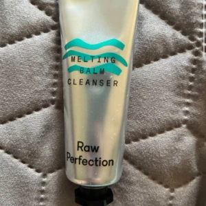Melting Balm Cleanser Raw Perfection - Silvrig tub med svart lock innehållande Melting Balm Cleanser från Raw Perfection. Rengöringsbalm som smälter bort smink och orenheter, perfekt för daglig ansiktsrengöring. Tuben har turkosa vågdetaljer och modern design.