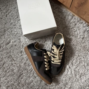 Maison Margiela GAT’s Black/Amber - Snygga svarta  sneakers från Maison Margiela med beige snörning och bruna gummisulor. Skorna har en mix av skinn och mocka, klassisk rund tå och retrovibbar. Perfekta för dig som gillar stilrena och tidlösa sneakers med en twist. Jag har en bild på kvittot sedan förra ägaren. Säljer pga att dem är för små! Byten är intressant!