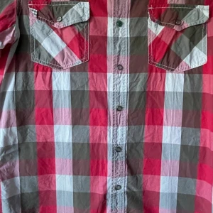 Rutig kortärmad skjorta City of Angels - Kortärmad skjorta från City of Angels i rött, grått och vitt rutigt mönster. Skjortan har två bröstfickor med lock och tryckknappar samt vit broderad logga ovanför ena fickan. Perfekt för dig som gillar en avslappnad och cool stil.