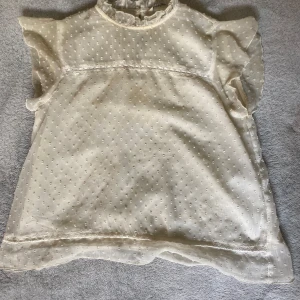 Vit blus med volangärm och prickar - Söt vit blus med små prickar och volangärmar. Blusen har en hög hals med rysch och knapp i nacken. Tunn och luftig, perfekt för vår och sommar. Ger en romantisk och trendig vibe.