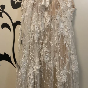 Långklänning med blommig spets och pärlor - Magisk långklänning i champagnefärgad mesh med vita 3D-blommor, pärlor och glittriga detaljer. Klänningen har v-ringning, dragkedja i ryggen och transparent lager över en beige bas. Perfekt för dig som vill sticka ut med en romantisk och lyxig look.