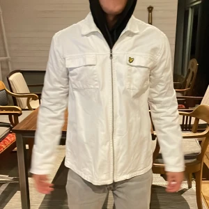 Vit overshirt från Lyle & Scott - Snygg vit overshirt från Lyle & Scott med dragkedja framtill och två bröstfickor. Jackan har klassisk krage och ikoniska Lyle & Scott-loggan på bröstet. Perfekt för dig som vill ha en stilren och clean look. Materialet känns lätt och smidigt. Perfekt för sommaren 