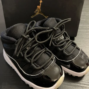 Jordan 11, svarta sneakers barnstorlek 22 - Snygga svarta Air Jordan 11 sneakers för barn med vit sula och glansiga detaljer i lack. Ovandelen är i mesh och syntet, med klassisk snörning och tydlig Jumpman-logga. Sulan har coolt JORDAN-mönster undertill. Perfekta för små sneakerheads! Tillhörande skokartong medföljer.
