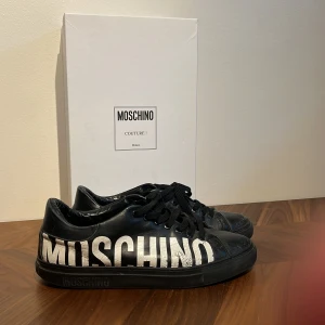 Svarta sneakers från Moschino - Coola svarta sneakers från Moschino med stor vit logga längs sidan. Skorna har snörning, rund tå och är tillverkade i skinn. Perfekta för dig som vill sticka ut med en statement-sneaker och gillar streetstyle. Priset är inte hugget i sten. 