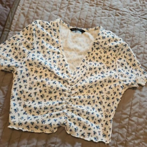 Vit blommig croppad topp från SHEIN - Superfin croppad topp från SHEIN i vit med små blå blommor. Toppen har v-ringning, korta ärmar och rynkdetalj framtill. Perfekt för varma dagar och ger en söt, somrig vibe.