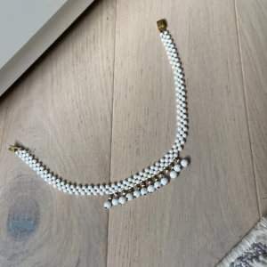 Vintage vitt pärlhalsband med gulddetaljer - Unikt vintage halsband med tre rader vita pärlor och små guldfärgade detaljer. Pärlorna är runda och i olika storlekar, vilket ger halsbandet en cool och klassisk look. Låset är dekorerat med ett vackert mönster i guldton. Perfekt för dig som gillar retrostil. 