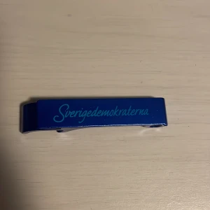 Blå kapsylöppnare med texten Sverigedemokraterna - Metallisk blå kapsylöppnare med texten 'Sverigedemokraterna' ingraverad på ovansidan. Kapsylöppnaren har en stilren och enkel design, perfekt för att öppna drickor med.Funkar perfekt till att ha som nyckelring!
