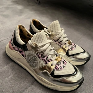 Roberto Cavalli sneakers med metallic storlek 36 - Unika Roberto Cavalli sneakers med vit mesh, silvriga och guldiga metallicdetaljer samt rosa och svart leopardmönster. Skorna har snörning och en chunky sula. Insidan är fodrad med leopardmönstrat tyg och framtill finns en guldfärgad logga. Ord.pris. 2800kr