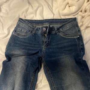Blå bootcut jeans med broderade fickor - Säljer ett par blå bootcut jeans med snygg slitning och kontrastsömmar. Baksidan har broderade fickor med lock och silvriga knappar. Jeansen har klassisk femficksdesign och är tillverkade i denim med en skön passform.
