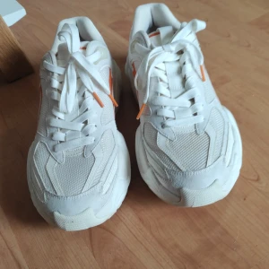 Vita chunky sneakers med orange detaljer - Säljer ett par vita chunky sneakers med coola orange detaljer på sidorna och hälen. Skorna har snörning och en rejäl, sportig sula. Ovandelen är i mesh och syntetmaterial för en fräsch look. Perfekta för dig som gillar streetstyle och vill sticka ut lite extra.