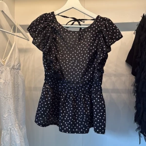 Svart prickig blus med volang - Supersöt svart blus med vita prickar och volangdetaljer längs axlarna. Toppen har en öppen rygg med knytband i nacken och en lätt utsvängd peplum nedtill. Perfekt för dig som gillar en lekfull och trendig look.