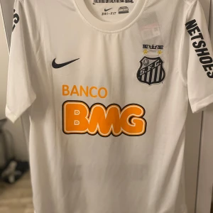 Santos Neymar Jr fotbollströja Nike L - Vit Santos fotbollströja från Nike med Neymar Jr och nummer 11 på ryggen. Tröjan har korta ärmar, svarta tryck och orange BMG-logga på bröstet. Tillverkad i lätt Dri-Fit material som andas. Snygg klubbsköld och sponsortryck på ärmarna. Den är helt ny det ända är att jag beställde i fel storlek.