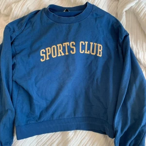 Blå sweatshirt från Cubus XS - Blå sweatshirt med gul text 'SPORTS CLUB' framtill. Klassisk rund halsringning och långa ärmar. Har några tecken på användning, se bild tre.