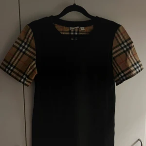Burberry t-shirt med rutiga ärmar - Svart t-shirt från Burberry med klassiska rutiga korta ärmar i beige, svart och rött. Gjord i 100% bomull för en skön känsla. Perfekt för dig som vill ha en stilren look med en touch av lyx och streetstyle.