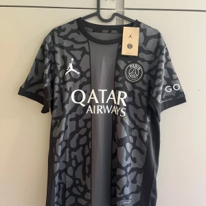 PSG x Jordan svart camo fotbollströja - Snygg svart och grå PSG fotbollströja i samarbete med Jordan. Tröjan har korta ärmar, camo-mönster och tryck med Qatar Airways på bröstet. Materialet är lätt och andas, perfekt för match eller träning. PSG-logga och Jumpman-logga på bröstet.