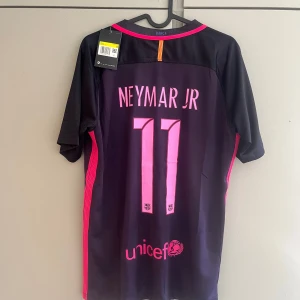 FC Barcelona Neymar Jr Nike tröja - Säljer en officiell FC Barcelona fotbollströja från Nike med Neymar Jr och nummer 11 på ryggen. Tröjan är mörklila med rosa detaljer, klubbmärke på bröstet och Qatar Airways-tryck. Tillverkad i lätt och ventilerande material, perfekt för dig som älskar Barca och Neymar.