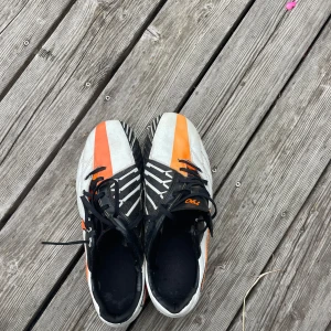 Nike fotbollsskor i svart, vitt och orange - Snygga fotbollsskor från Nike med svart, vitt och orange färg. Skorna har snörning, metall-dobbar och ett grafiskt mönster på ovandelen. Perfekta för dig som vill sticka ut på planen med stilren design och bra grepp.