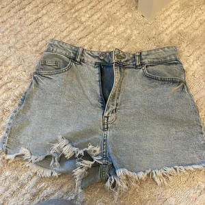 Ljusblå jeansshorts med fransig kant - Säljer ett par ljusblå jeansshorts med rå och fransig kant nertill. Klassisk femficksmodell med bälteshällor och dragkedja fram. Perfekta för sommaren och ger en avslappnad vibe till din outfit.