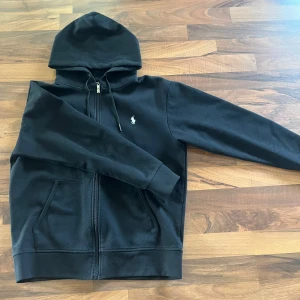 Svart hoodie från Polo Ralph Lauren - Svart hoodie från Polo Ralph Lauren med dragkedja, huva med snörning och klassisk vit logga på bröstet. Tröjan är tillverkad i mjuk bomull. Perfekt för en soft och stilren look.