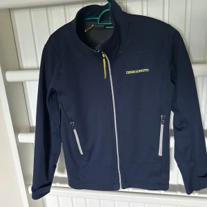 Mörkblå vindjacka strl 140 från Didriksons - Mörkblå vindjacka från Didriksons med hel dragkedja och två fickor framtill. Jackan har sportig look, diskret logga på bröstet och detaljer i gult och grått. Perfekt för blåsiga dagar och enkel att matcha med det mesta.