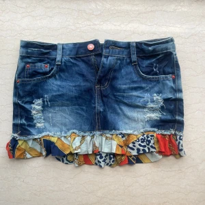 Jeanskjol med volang och mönster - Cool kort jeanskjol med slitna detaljer och färgglad volangkant i blått, rött, vitt och gult. Kjolen har klassiska fickor fram och bak samt bälteshällor. 