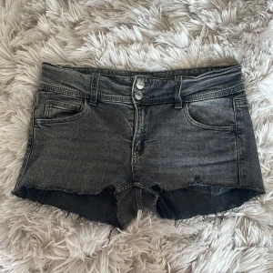 Mörkgrå jeansshorts från Gina Tricot - Snygga mörkgrå jeansshorts från Gina Tricot med råa kanter och dubbla knappar framtill. Klassisk femficksmodell med bakfickor som har knappdetaljer. Perfekta för sommardagar när du vill ha en cool och avslappnad look.