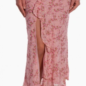 Rosa blommig kjol med volang från Neo Noir - Lång rosa kjol med blommigt mönster och volangdetalj längs omlottkanten, stl 34. Kjolen har en snygg slits framtill som ger en cool vibe. Perfekt för dig som gillar romantiska och somriga plagg med extra detaljer.