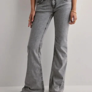 Grå bootcut jeans från Nelly - Snygga grå jeans från Nelly med bootcut passform och coola svarta broderade detaljer på bakfickorna. Jeansen har hög midja, klassisk femficksdesign och knäppning med tre knappar. Perfekta för dig som gillar en trendig och avslappnad stil.